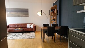A modern living room with a brown leather sofa, a dining table with chairs, a bookshelf, and a large painting on the wall.