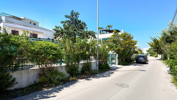 Una calle estrecha bordeada de edificios residenciales, un coche aparcado y abundante vegetación.