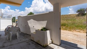 An outdoor patio with a white table and chair, a planter with green plants, and a view of a gravel path and hills. - 3463 Villetta Naomi by Barbarhouse (Porto Cesareo)