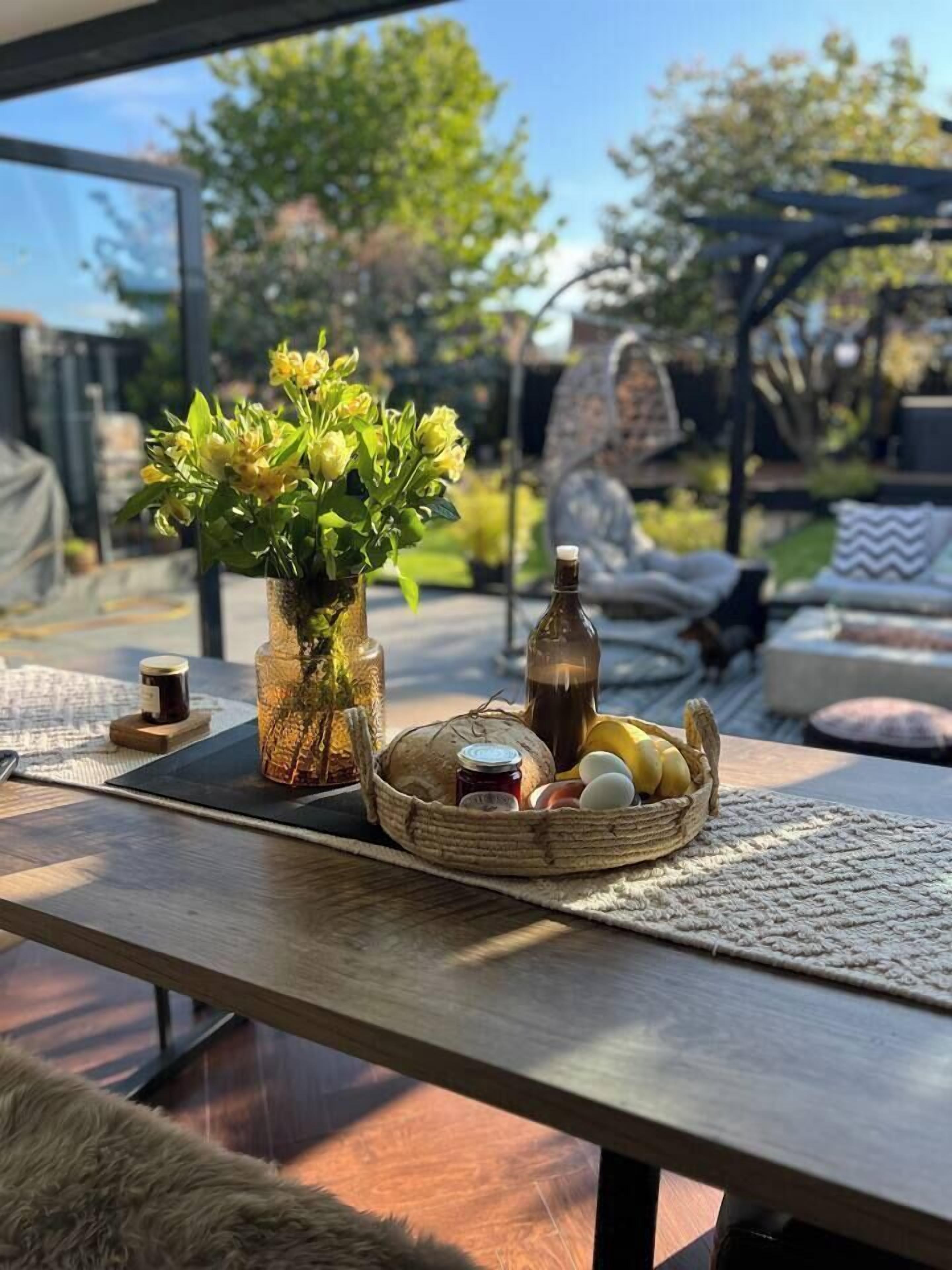 A wooden table with a vase of flowers, a bottle, and a basket of eggs and bread.