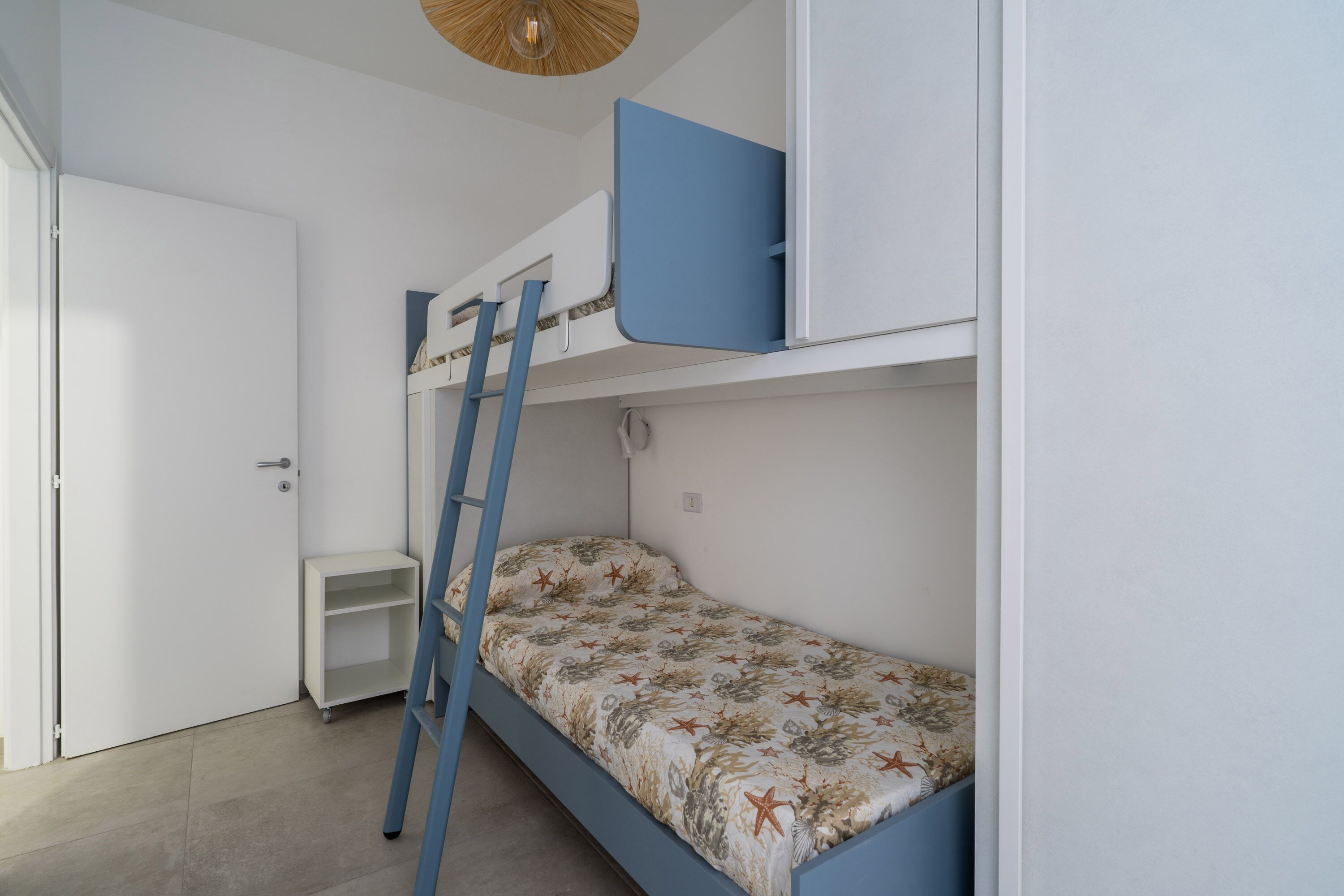 A bunk bed with a ladder, a small shelf, and a door in a room with white walls and a ceiling light.