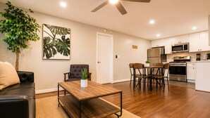Un salon moderne comprenant un canapé, une table basse et un coin repas avec des chaises. On y trouve également une cuisine équipée d’appareils électroménagers et de meubles de rangement.
