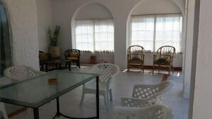 Zona de dinar amb taula de vidre i cadires blanques, finestres grans amb cortines i una planta.