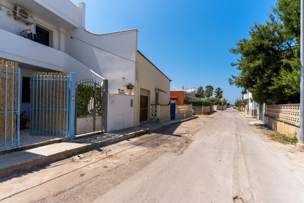 Une rue résidentielle tranquille, bordée de maisons blanches, avec un portail bleu et un ciel d’un bleu limpide.