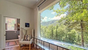 Une pièce dotée d’une grande fenêtre donnant sur une zone boisée, d’un fauteuil avec un coussin et d’un petit tapis sur le sol.