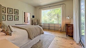 Une chambre à coucher avec un lit, un ventilateur de plafond, une grande fenêtre avec des rideaux et une commode en bois.