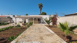 Una propiedad residencial con entrada pavimentada, porche cubierto y jardín con diversas plantas.