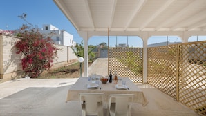 Un área de comedor al aire libre con una mesa dispuesta para dos personas, rodeada de sillas blancas, bajo una pérgola blanca.