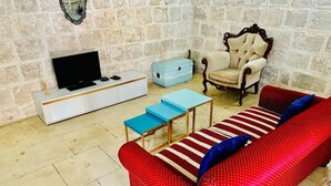 A living room with a stone wall, a television, a white cabinet, a blue chest of drawers, a red sofa with blue cushions, and a wooden chair. - Serenity 2  studio apartment with small terrace (Ħal Qormi)
