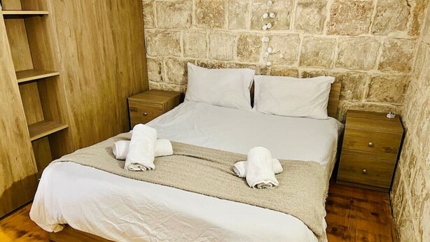 A neatly made bed with white linens and two rolled towels, wooden nightstands, a wooden wardrobe, and a stone wall background. - Serenity 2  studio apartment with small terrace (Ħal Qormi)
