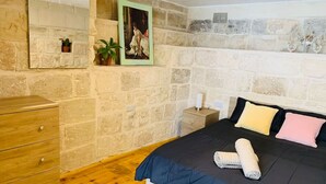 A bedroom with a stone wall, a wooden dresser, a bed with a dark blue bedspread and two pillows, and a framed picture on the wall. - Serenity 1 -cosy apartment in Qormi (Ħal Qormi)