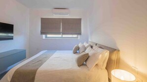 Een slaapkamer met een bed, een raam met jaloezieën, een wandtelevisie en een bedlampje.