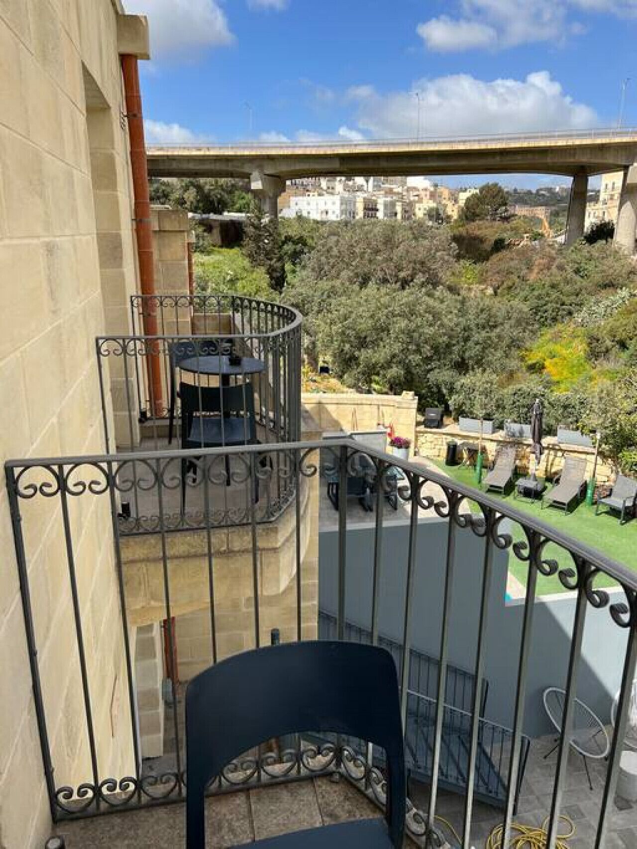 Un balcó amb barana corba, una taula petita i una cadira. Al fons, es veu un pont i un paisatge urbà amb edificis i vegetació.