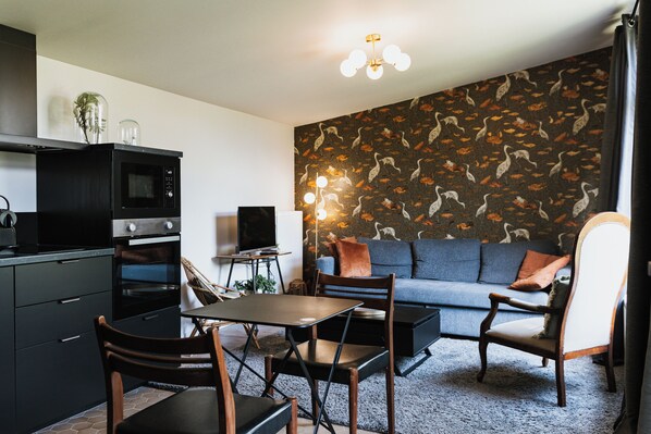 A modern kitchen with a microwave, oven, and dining area. There is a sofa and a television in the living area. - Les Glycines, Chambre Avec Jardin (Criel-sur-Mer)