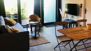 A modern living room with a sofa, a dining table, chairs, a TV, and a large window with a view. - Bel Air, Accès Piscine - Jacuzzi Privé - Terrasse (Criel-sur-Mer)