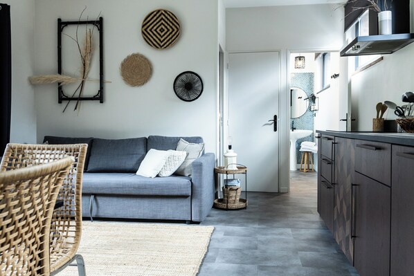 A modern living room with a grey sofa, wicker chair, and a small side table. A kitchen area is visible with dark cabinets and a window. - Appartement - Piscine - Terrasse, Beau Séjour (Criel-sur-Mer)