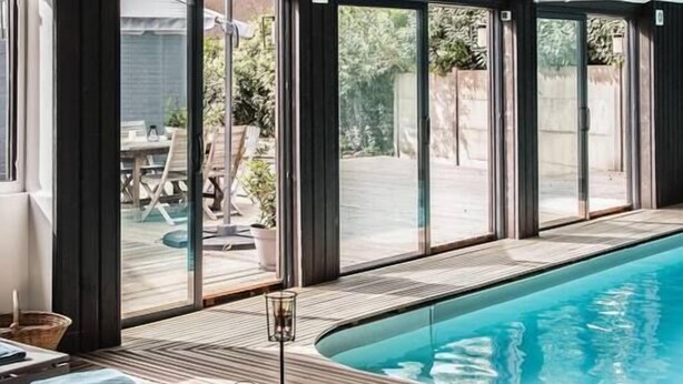 Un espace aménagé au bord de la piscine, avec une terrasse en bois, une piscine à moitié fermée par des parois de verre et une vue sur l’extérieur.