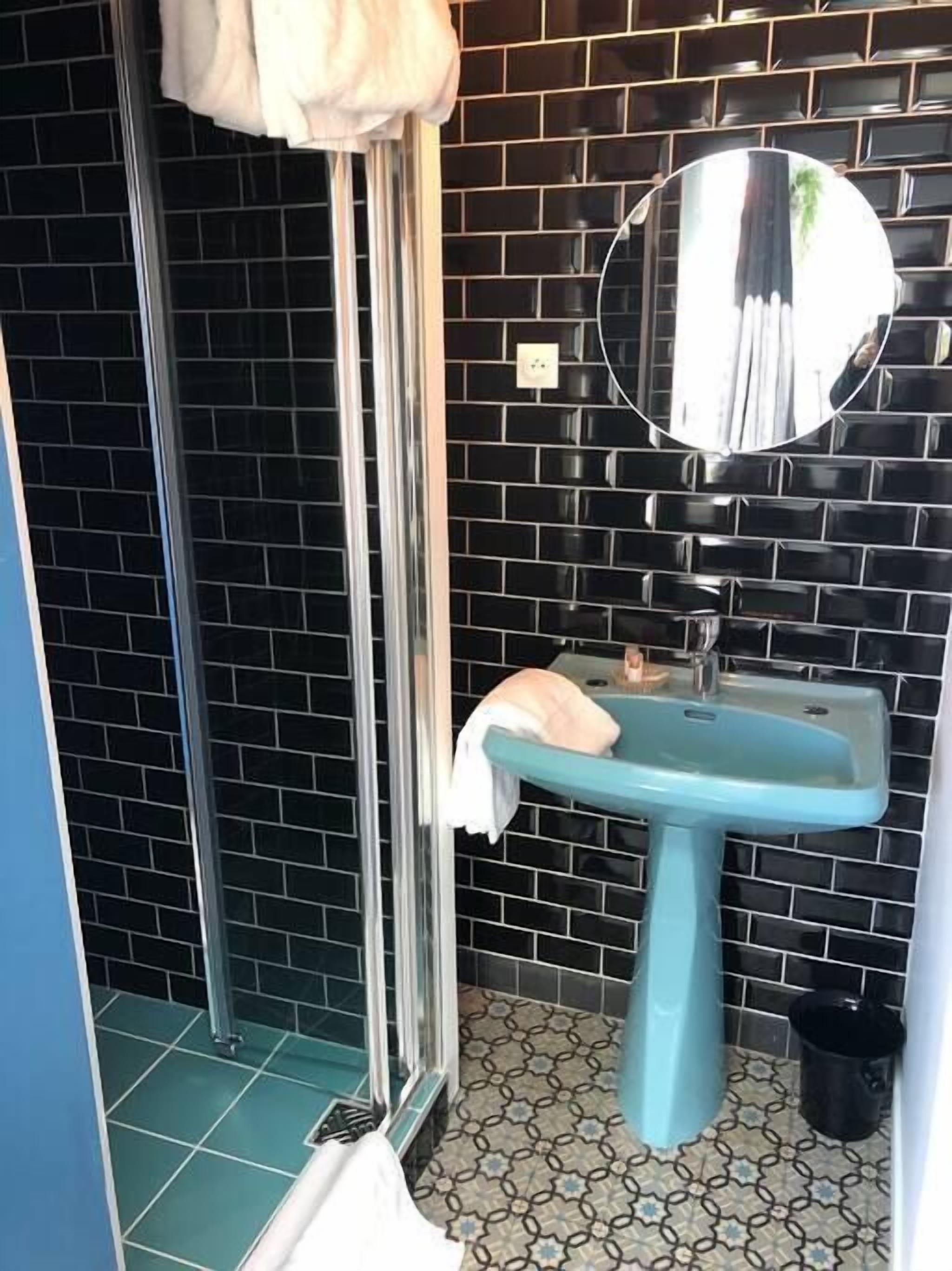 Une salle de bain avec un mur recouvert de carreaux noirs, un lavabo bleu, un miroir et un espace douche.