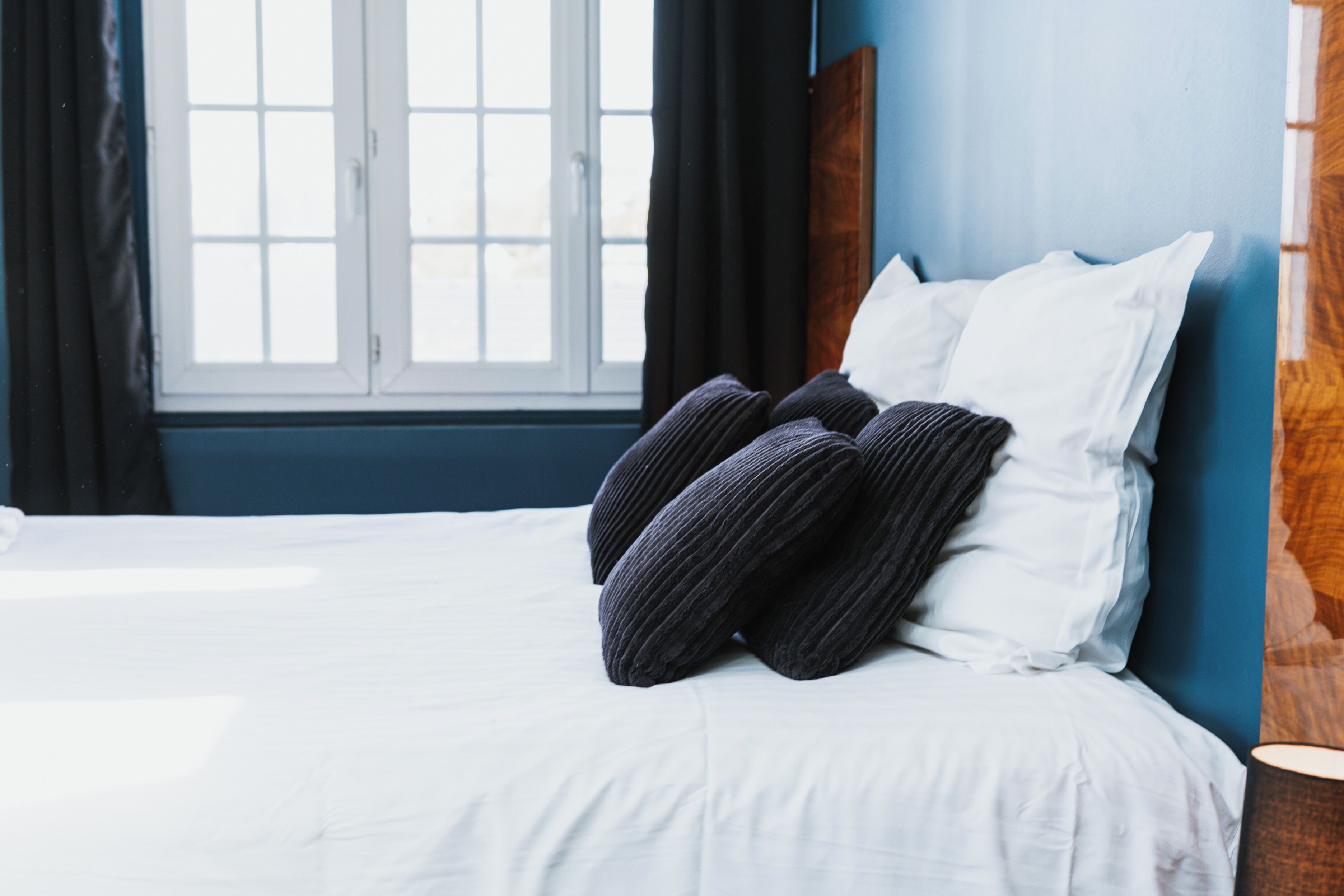 A bed with white bedding and dark pillows, a wooden headboard, and a window with curtains.
