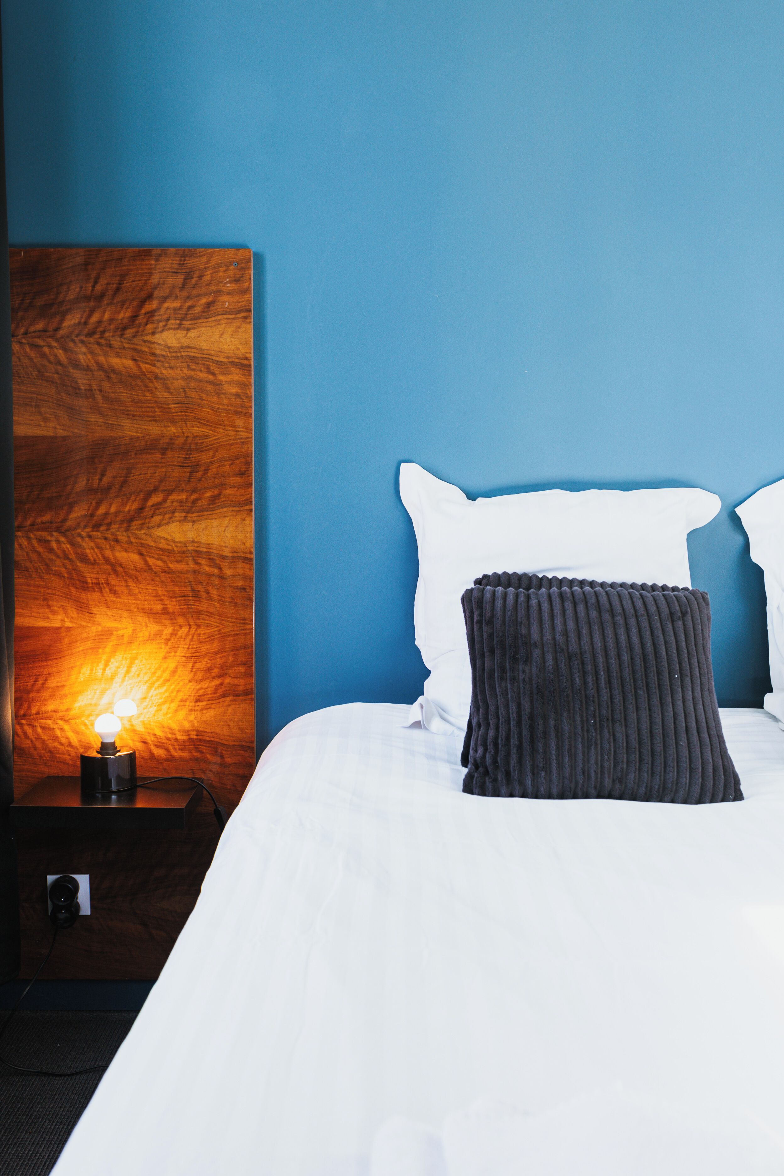 A neatly made bed with white bedding, a dark headboard, and a bedside table with a lamp.