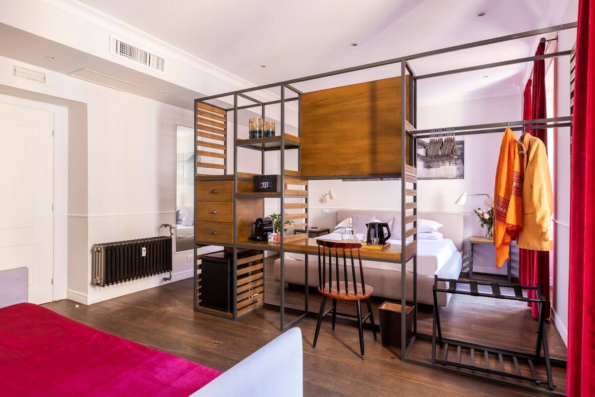 A modern, compact room with a Murphy bed, a small desk, and a chair. There is a radiator, a window with curtains, and a red throw blanket on the bed.