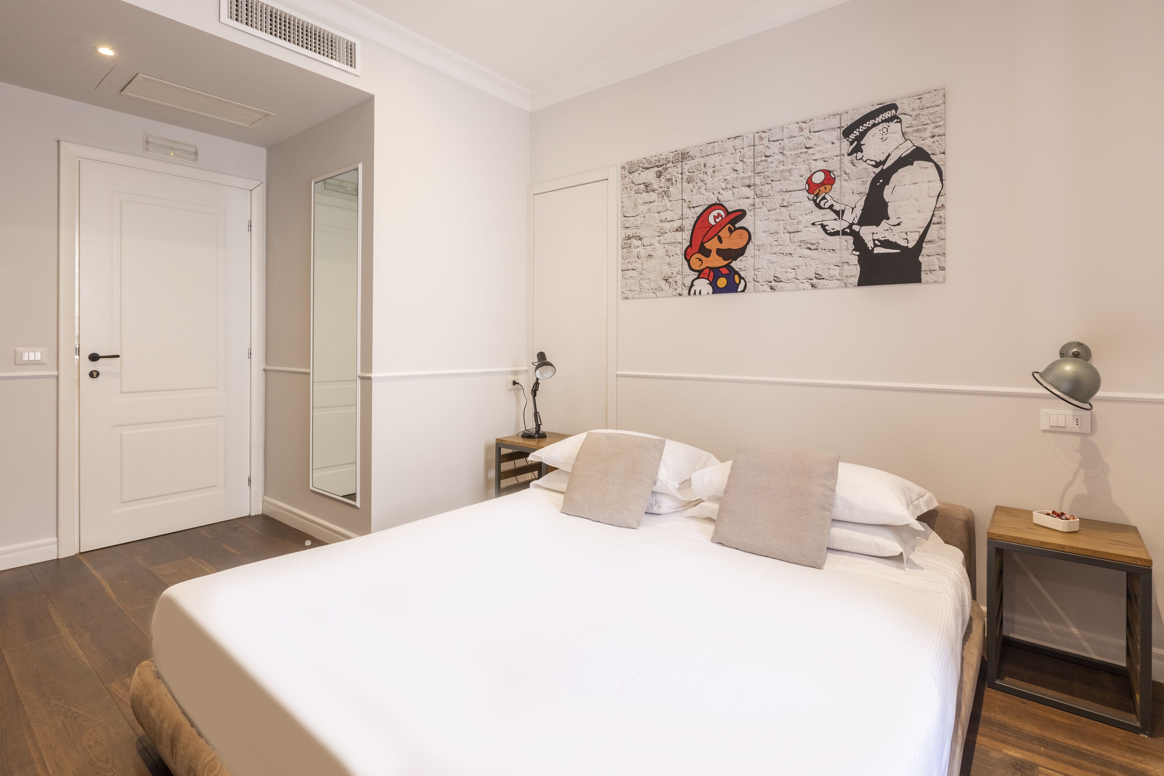 A bedroom with a large bed, a nightstand with a lamp, a wall-mounted artwork, and a door.