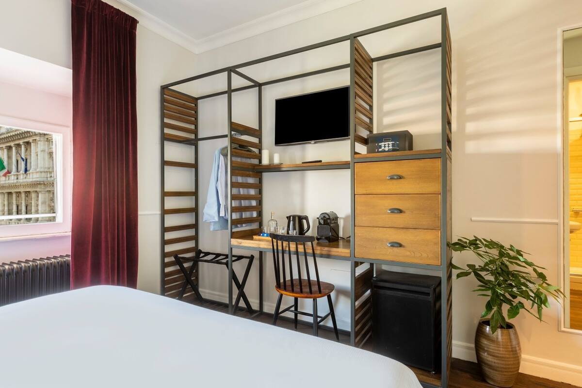 A hotel room with a bed, a desk with a chair, a TV, a coffee maker, and a plant.
