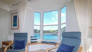 Ein Zimmer mit zwei blauen Polsterstühlen, einem kleinen Holztisch und Blick auf einen Hafen durch große Fenster.