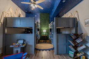 A room with bunk beds, a ceiling fan, a starry ceiling, a green wall, a desk with a lamp, and a bookshelf.