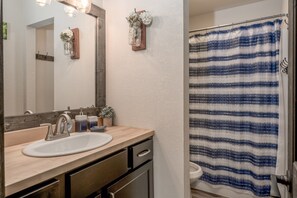 Une salle de bain avec un meuble-lavabo en bois, un lavabo blanc, un miroir, un rideau de douche et une petite plante en pot.