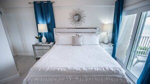 Una cama bien hecha con ropa de cama blanca y un borde con flecos, flanqueada por dos mesitas de noche con lámparas y jarrones decorativos. Un espejo redondo cuelga sobre la cama, y la habitación cuenta con un ventanal con cortinas azules.