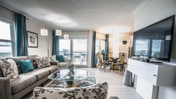 Una sala de estar moderna con sofá, sillón y mesa de centro, un comedor con sillas y un televisor sobre un mueble.