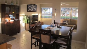 A modern dining area with a large table, chairs, a TV, and a kitchen area with cabinets and a plant. - Fabulous one Bedroom in West Bay (West Bay)