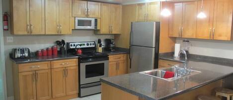 A kitchen with wooden cabinets, a microwave, a refrigerator, a stove, and a sink. There are stools near the sink and a trash bin in the corner.