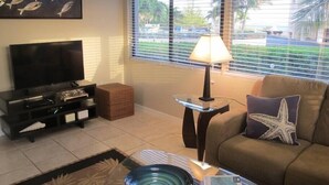 A living room with a TV, a glass coffee table, a lamp, a sofa with a starfish pillow, and a painting of fish on the wall. - Fabulous one Bedroom in West Bay (West Bay)