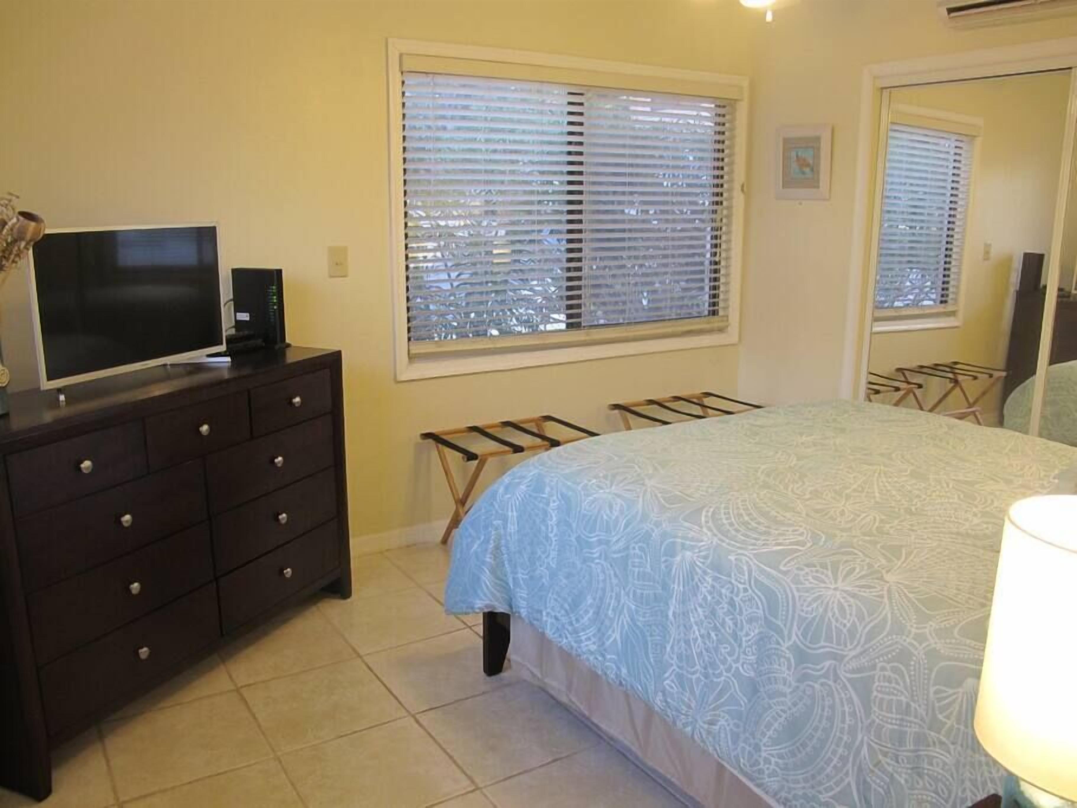 A bedroom with a bed, dresser, TV, and window with blinds.