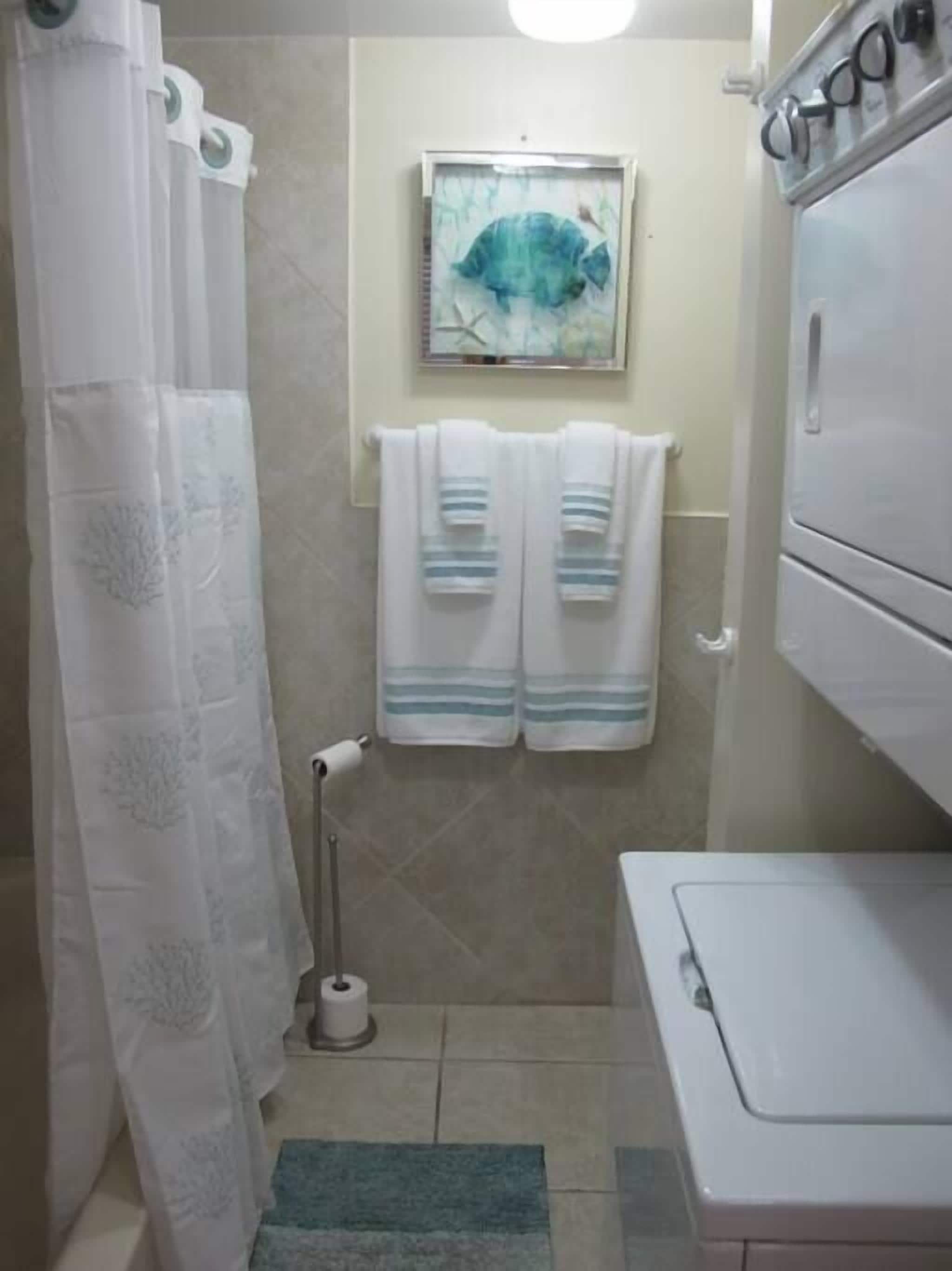 A bathroom with a shower curtain, a framed artwork on the wall, two towels hanging on a rack, a toilet paper holder, and a washing machine.