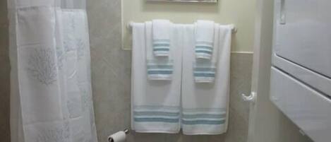 A bathroom with a shower curtain, a framed artwork on the wall, two towels hanging on a rack, a toilet paper holder, and a washing machine.