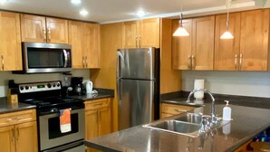 A modern kitchen with wooden cabinets, stainless steel appliances, a central island with stools, and a tiled floor. - Fabulous one Bedroom in West Bay (West Bay)