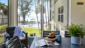 Ein Balkon mit Blick auf einen Pool, Palmen und einen Strand. Dort stehen ein Stuhl, ein Tisch mit einer Zeitung, ein Hut, eine Dose und ein Blumentopf.