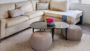 Ein modernes Wohnzimmer mit einem beigefarbenen Ecksofa, zwei runden Hocker, einem Glastisch mit Blumenvase, einem Deckenventilator und Kunstwerken an der Wand.