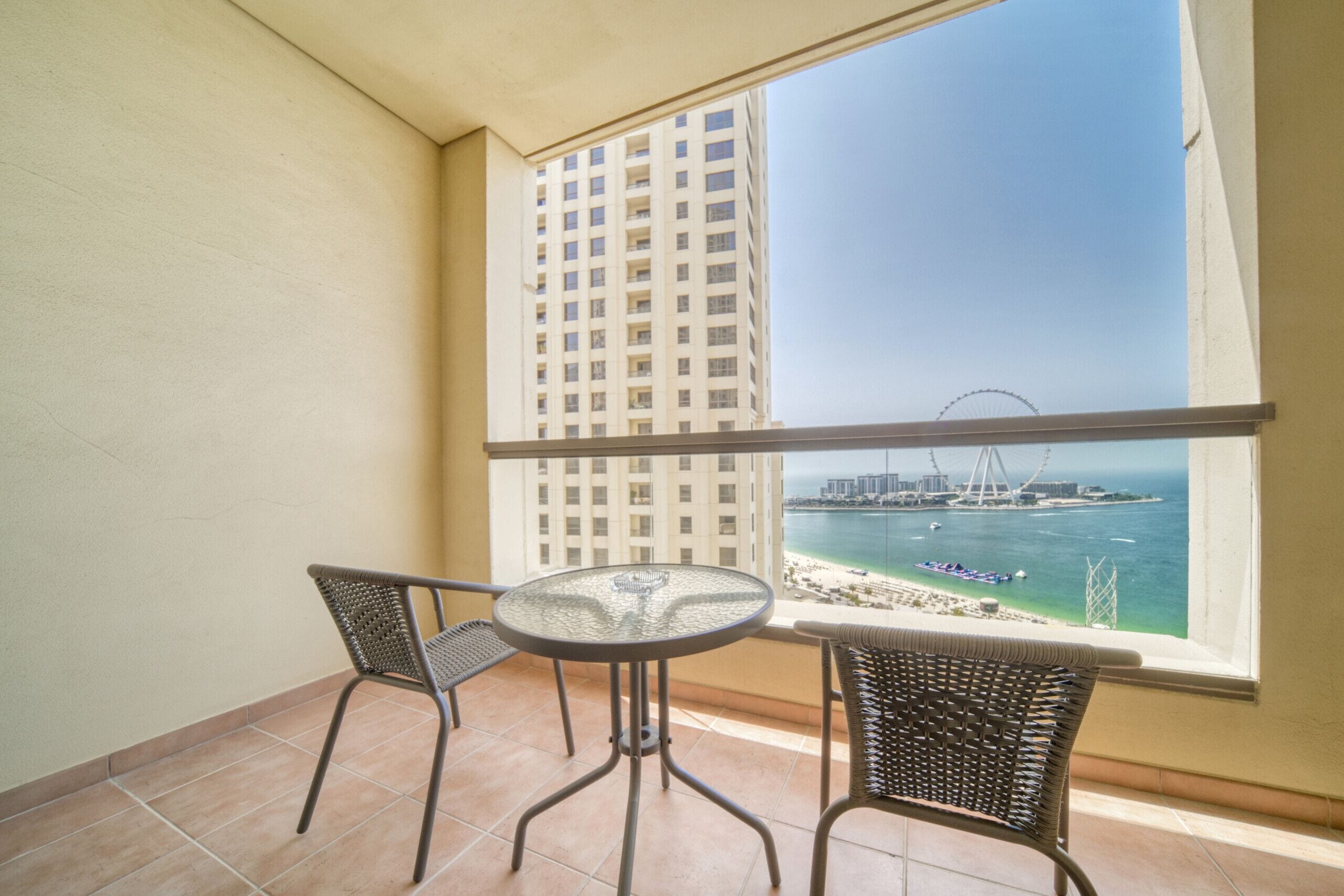 A balcony with two wicker chairs and a glass table, overlooking a beach and a Ferris wheel.