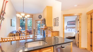 Una cocina con encimera de granito, armarios de madera y lavavajillas. Hay una zona de comedor con sillas y mesa de madera. Se ve una cama al fondo.