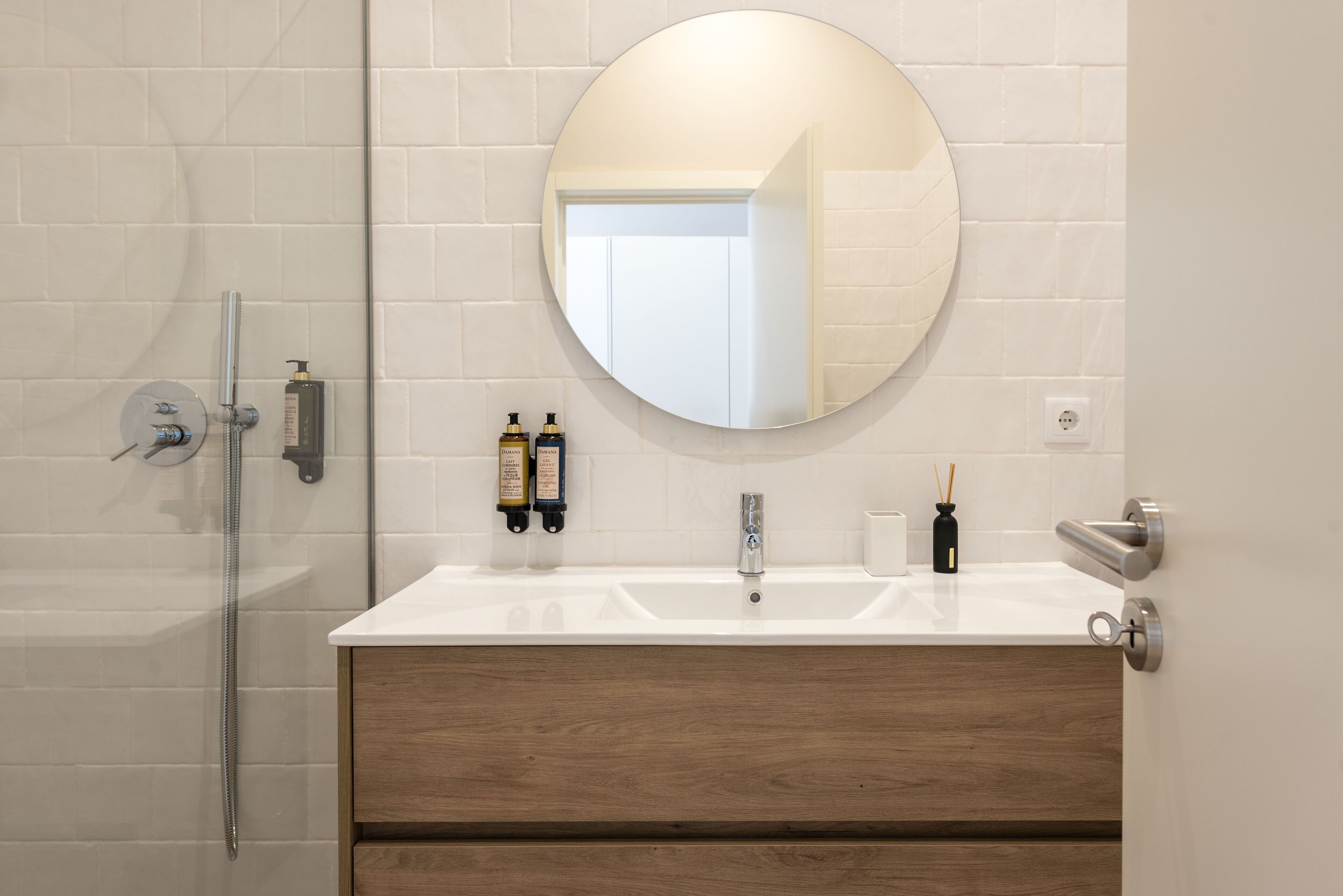 Een moderne badkamer met een douchecabine van glas, een ronde spiegel en een houten wastafelmeubel met twee wastafels.