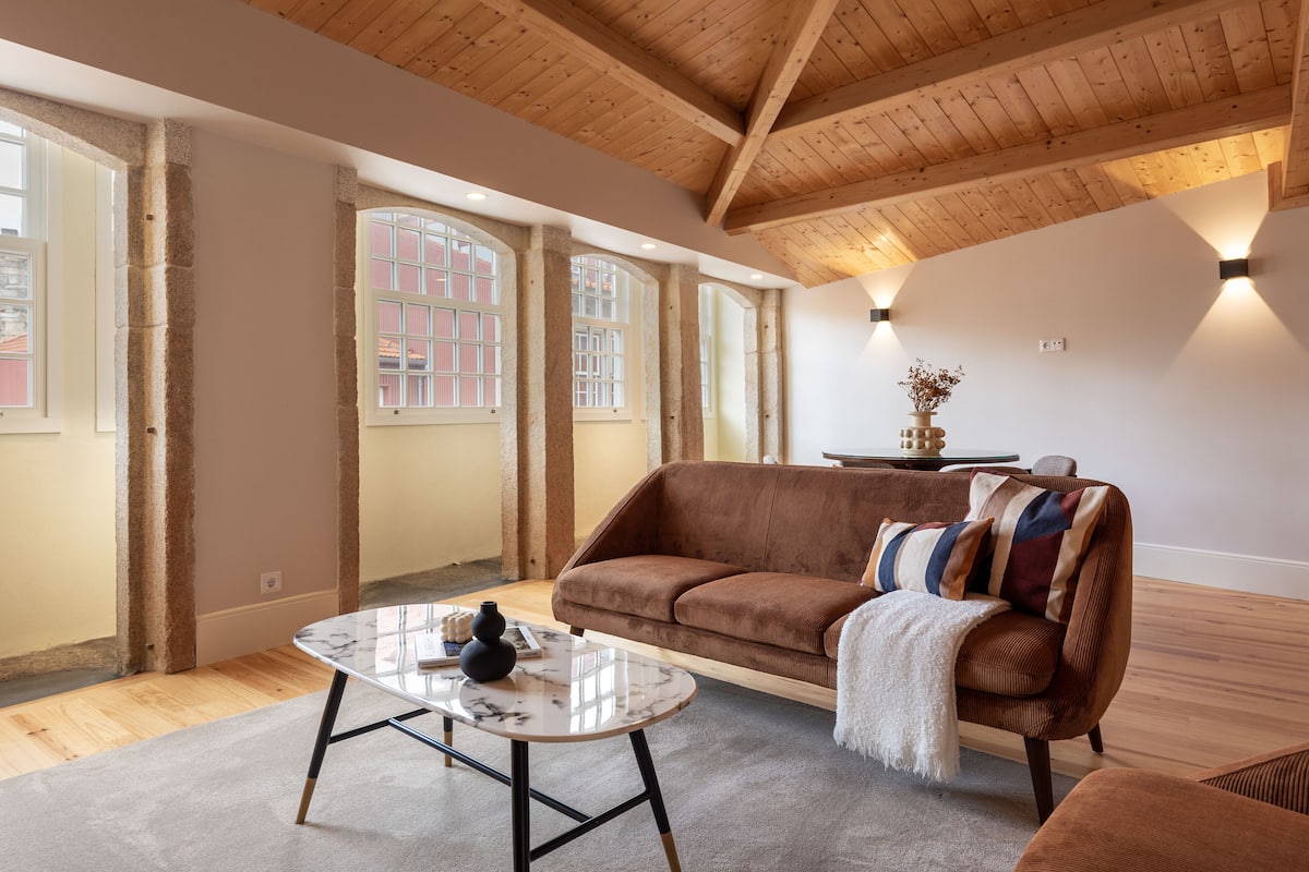A spacious living room with a high wooden ceiling, large windows, a brown sofa, and a marble coffee table.