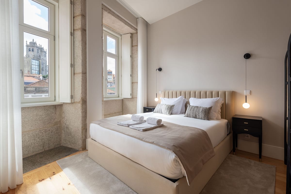 A bedroom with a large bed, two bedside tables, a window with a view of a building, and a modern light fixture.