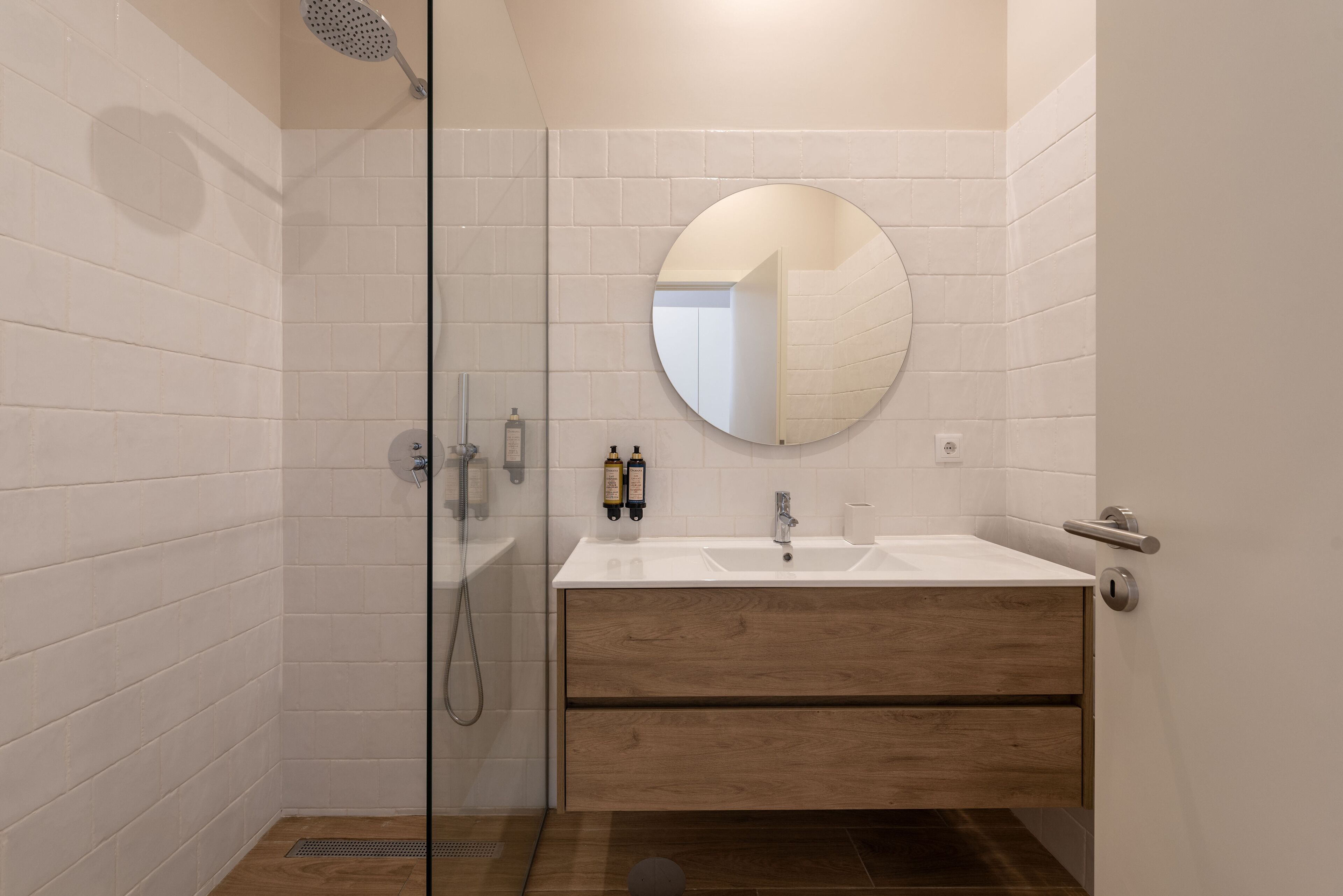 Un baño moderno con cabina de ducha de vidrio, tocador de madera con espejo redondo y pared de azulejos blancos.