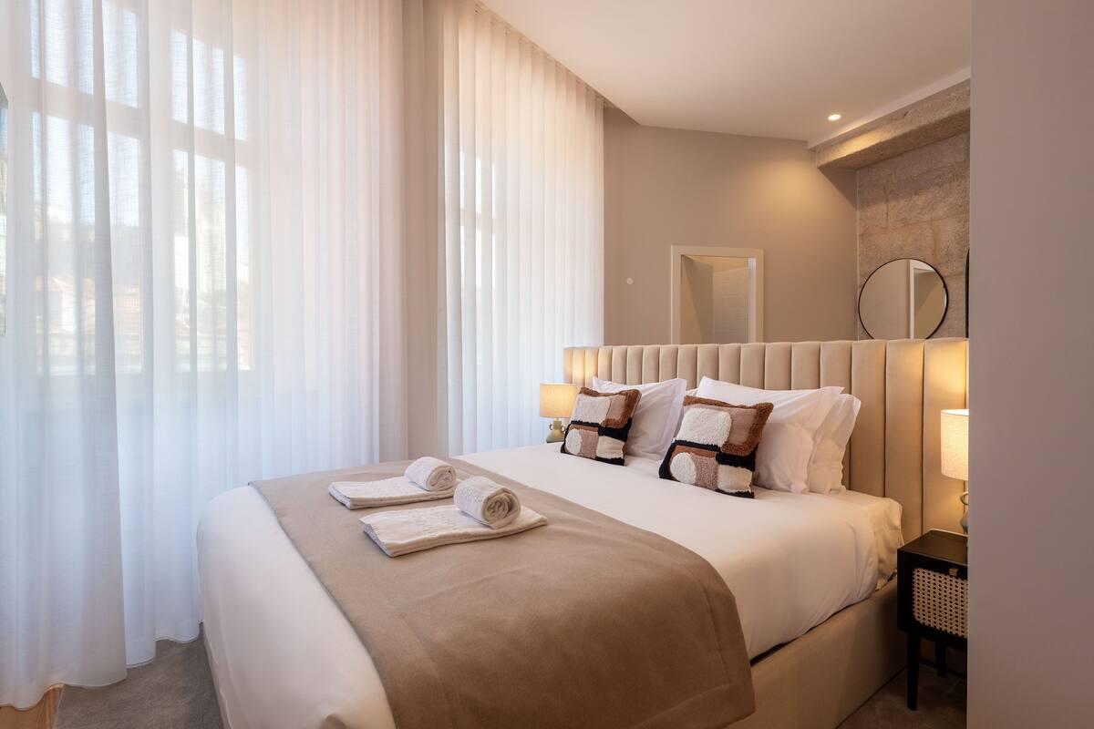 A neatly made bed with a beige bedspread and pillows, a bedside lamp, and a wall-mounted mirror.