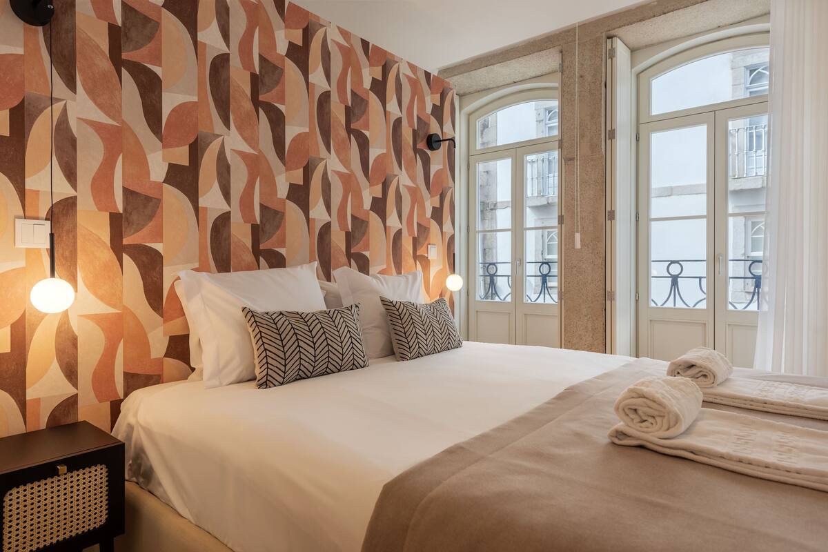 A neatly made bed with white and patterned pillows, a beige blanket, and two rolled towels.