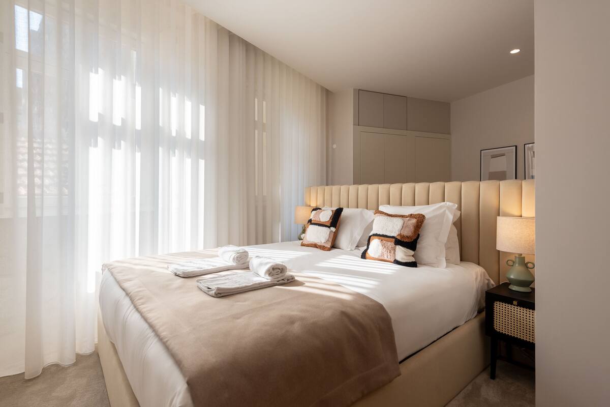 A modern hotel room with a large bed, a bedside table with a lamp, and sheer curtains.